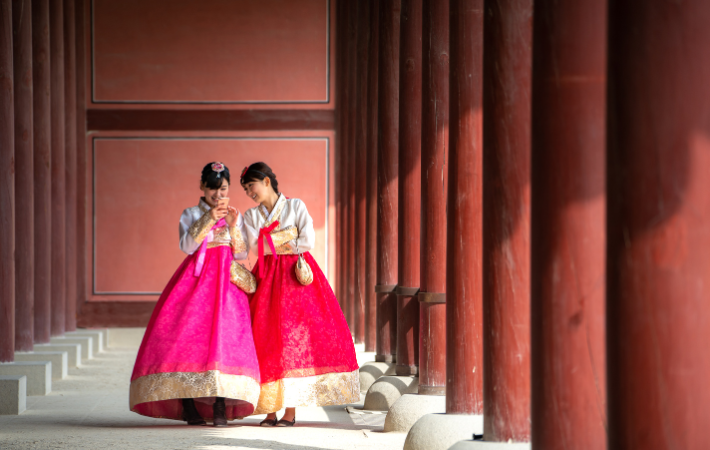 Duas jovens andando pelo Palácio vestidas em trajes tradicionais e rindo enquando olham o celular que uma tem em mãos
